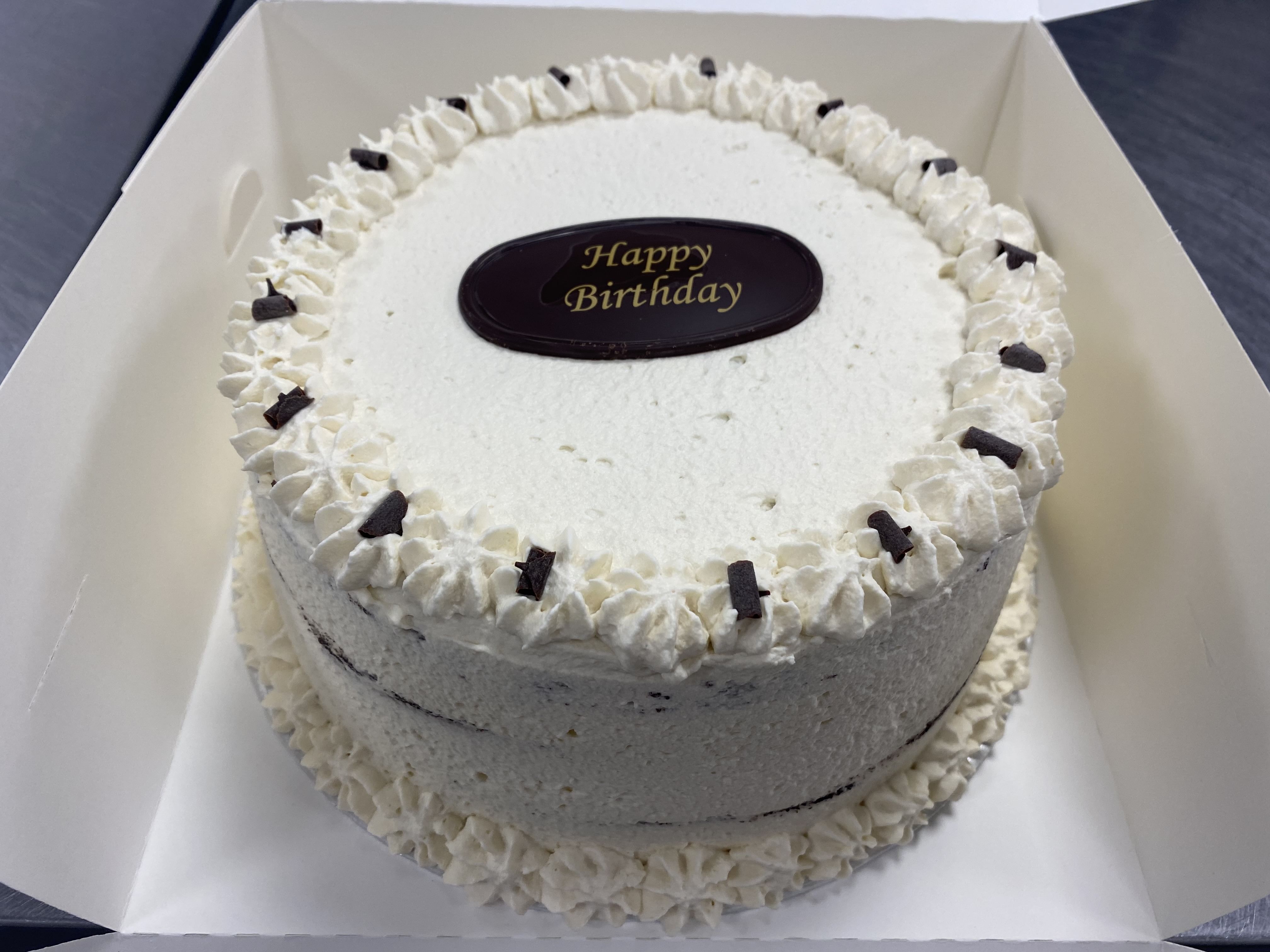 A round, white-frosted birthday cake resembling a Black Forest cake is inside an open white box. Decorated with small, piped white rosettes along the top edge and base, it features chocolate goodness in the form of small pieces along the top border. A chocolate plaque in the center reads 'Happy Birthday.'